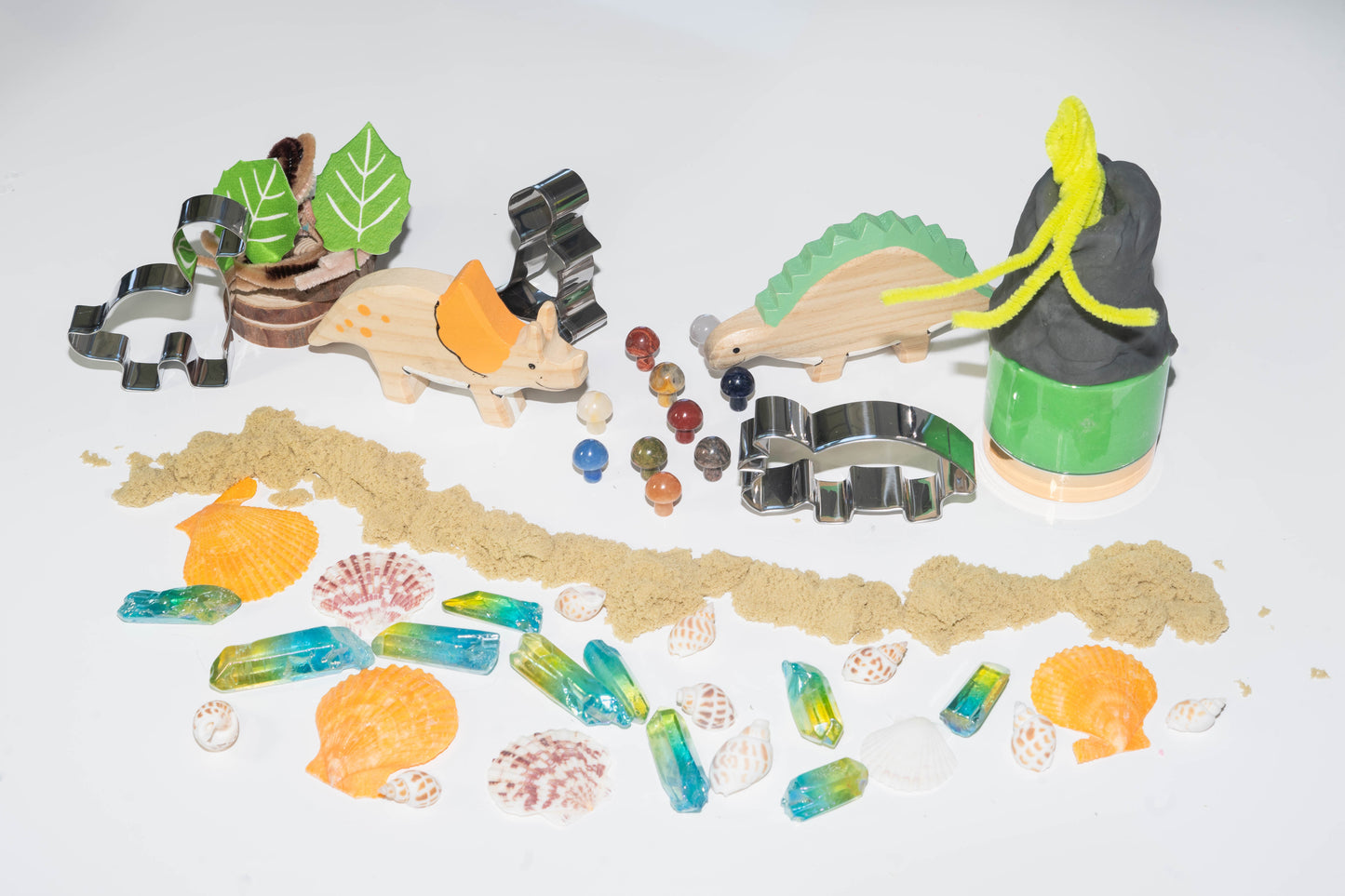 2 wooden dinosaurs, 2 metal dinosaur cookie cutters, dough, sand, and green and yellow ombre crystals, with sea shells and leaves made of seed paper