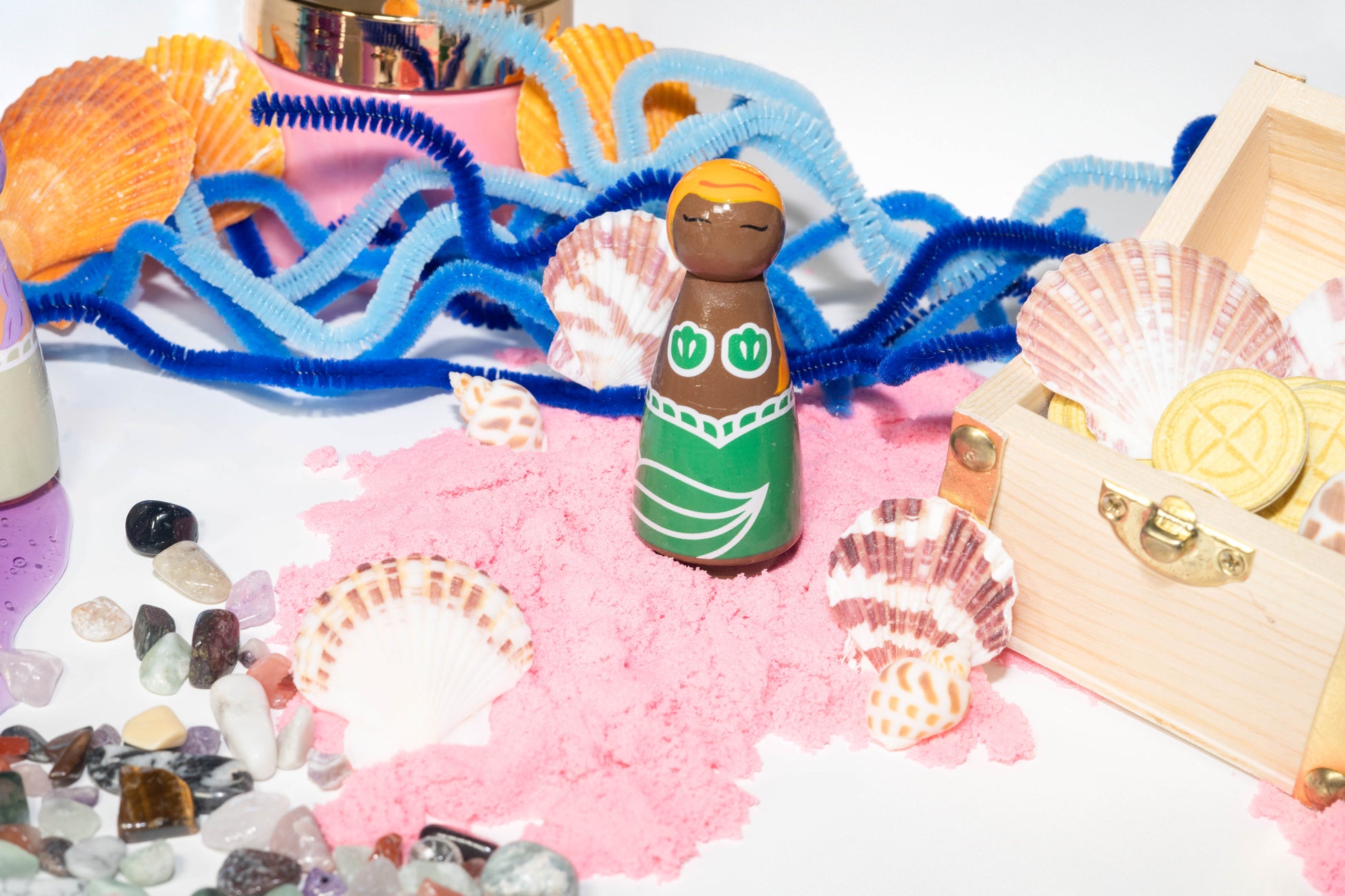 Wooden mermaid peg doll hand painted with green tail and matching top set in a scene with pink kinetic sand, wooden treasure chest, seed paper coins, and blue pipe cleaners