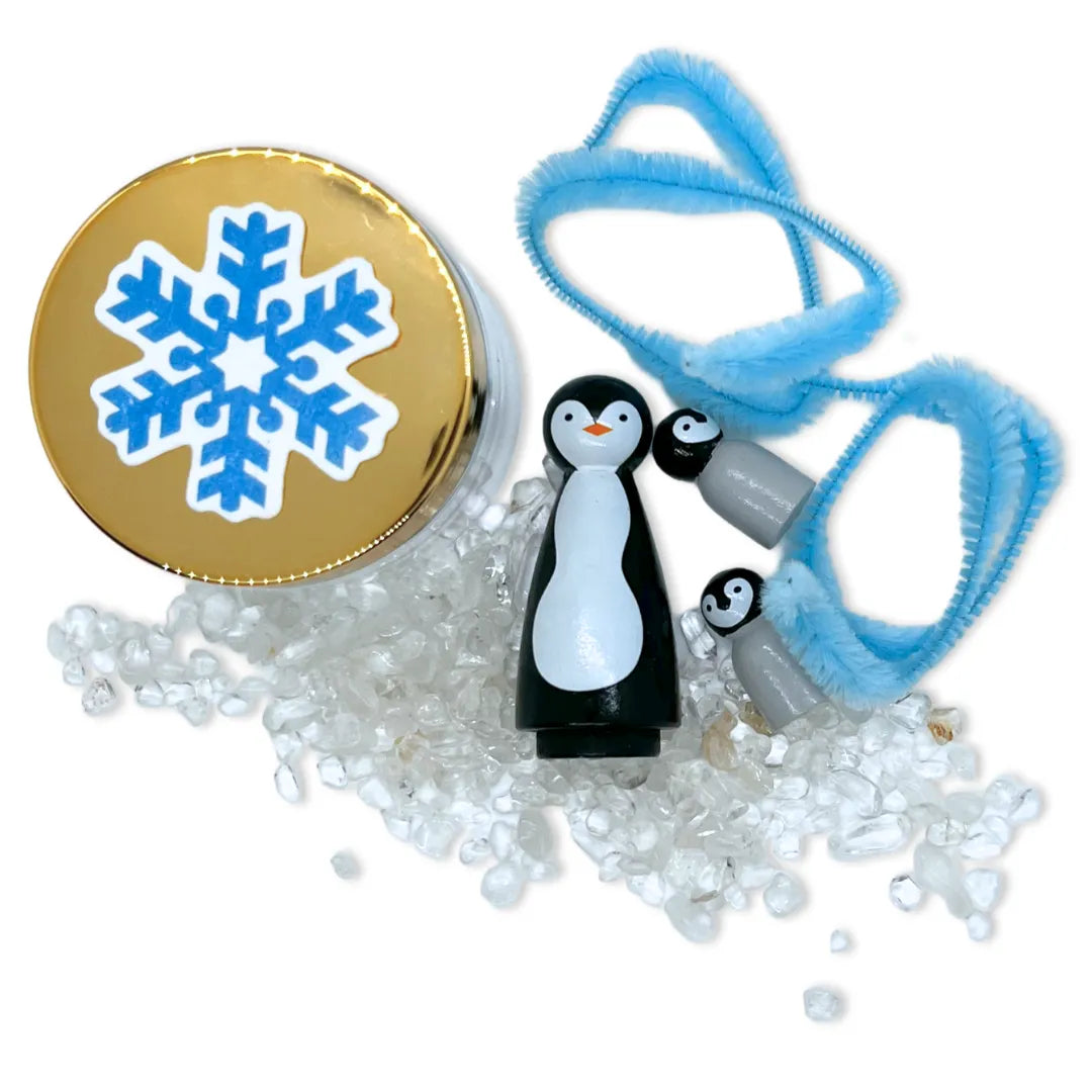 A Penguin dad peg doll, a pair of baby penguin peg dolls on top of a field of clear crystals along with ice blue pipe cleaners, a jar of white play dough and a snow flake shaped wild flower snowflake.